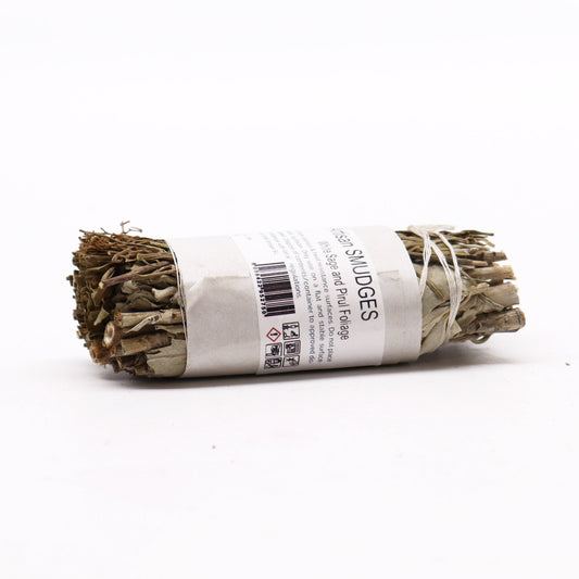 A sage smudge stick rests on a white surface. The stick is bundled and tied with twine, labeled "Artisan SMUDGES White Sage and Pirul Foliage". Further instructions are included on the label.