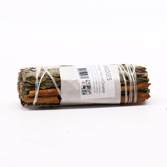 A smudge stick, composed of sage, cedar, and cinnamon, is bound with twine. It rests on a white surface. The label reads: "SMUDGES White Sage, Cedar and Cinnamon Only use on a flat and stable surface heat resistance surfaces. Do not place of contents/container to approved dis- local regulations 056422952774" Several pictograms are also present on the label.