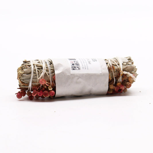 A sage smudge stick, bound with twine and adorned with pink peppercorns, rests on a white surface. The label reads: GES Pink Pepper Smudge. Warnings: Do not place near a flammable surface. Burn on approved dish.