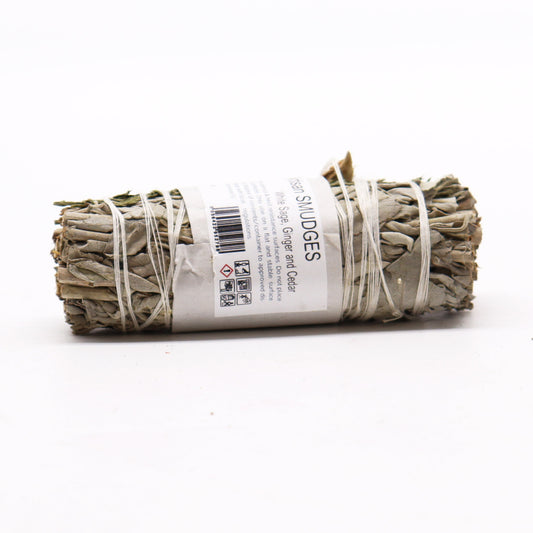 A smudge stick of white sage, ginger, and cedar rests on a white surface. It is bound with twine and labeled with product information.