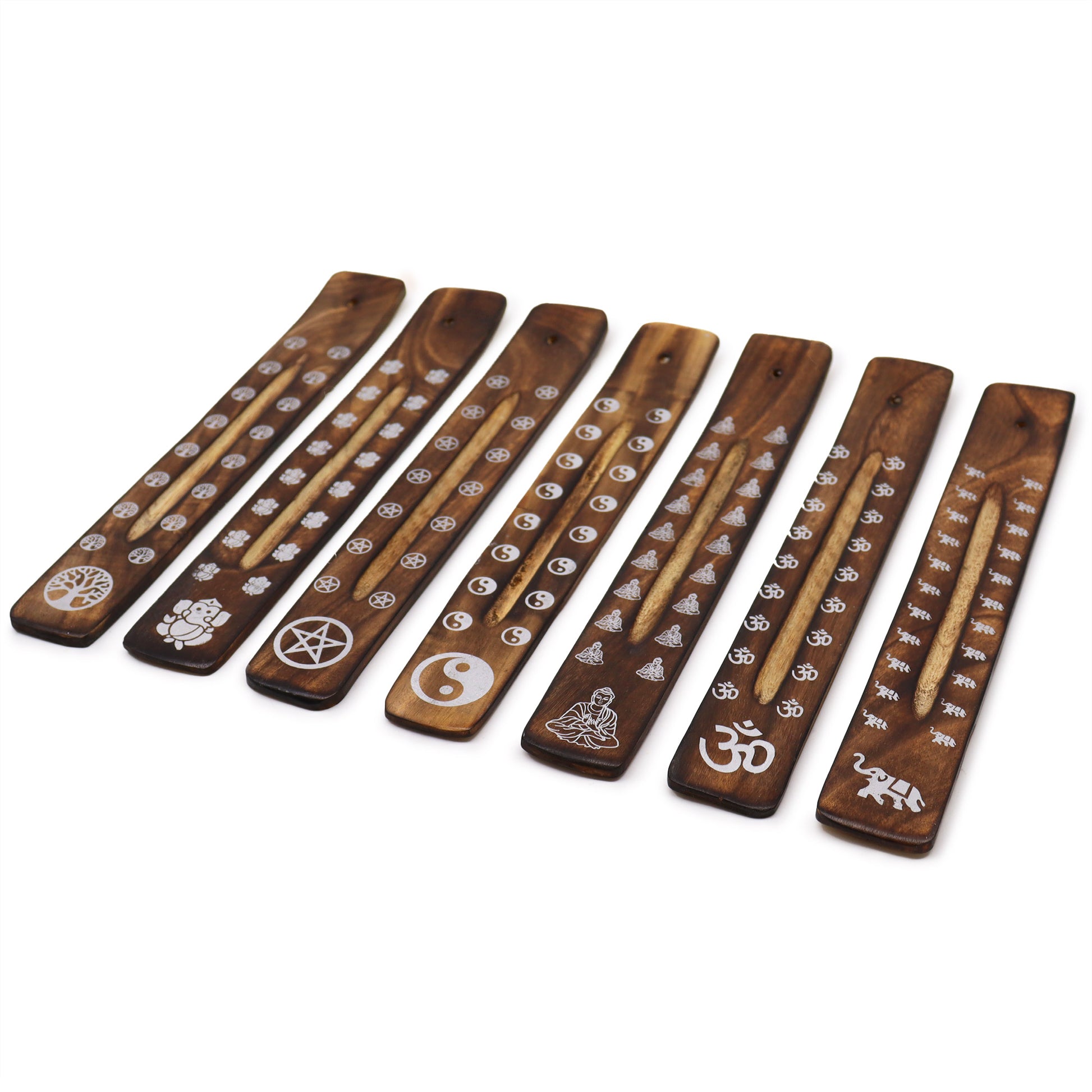 Wooden incense holders with engraved symbols, including tree of life, Buddha, pentacle, yin-yang, seated figure, Om, and elephant, are lined up, displaying detailed decoration on a white surface.