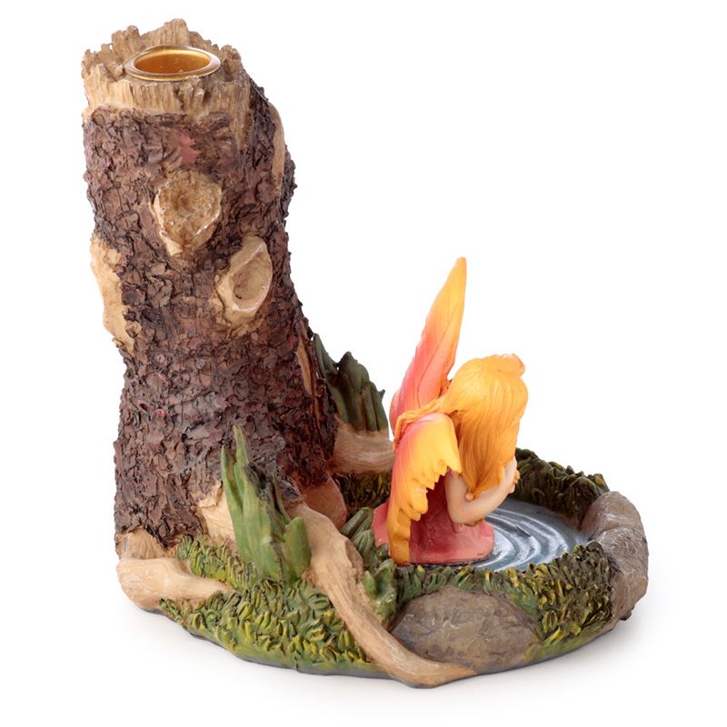 A small fairy with orange wings kneels by a circular pond, set against a tall, textured tree trunk with a candle holder, surrounded by green foliage.