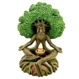 A tree-like figurine with a human face sits cross-legged, holding a small candle. Its leafy canopy forms hair, and its roots spread outward on a flat base.