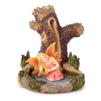 A figurine fairy with orange wings and a pink dress kneels beside a tree stump, surrounded by greenery and water on a decorative base.