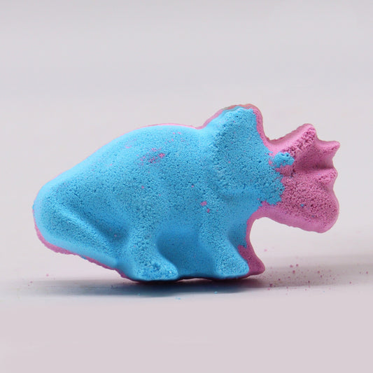 A blue and pink dinosaur-shaped bath bomb rests on a light surface, with some powder scattered around, creating a colorful and playful appearance.