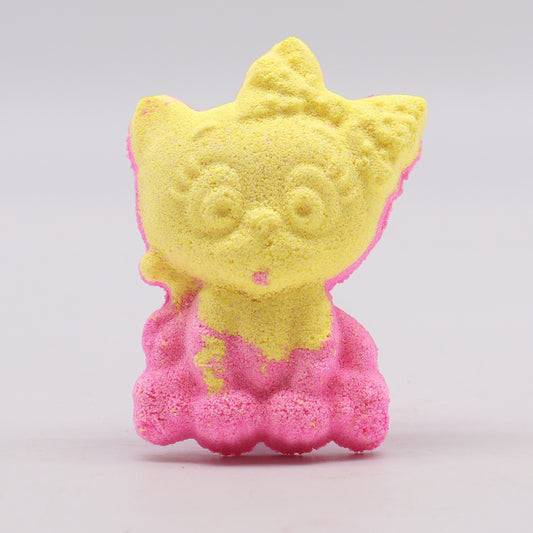 A yellow and pink cat-shaped bath bomb sits upright on a light gray surface, featuring a detailed face and playful expression.