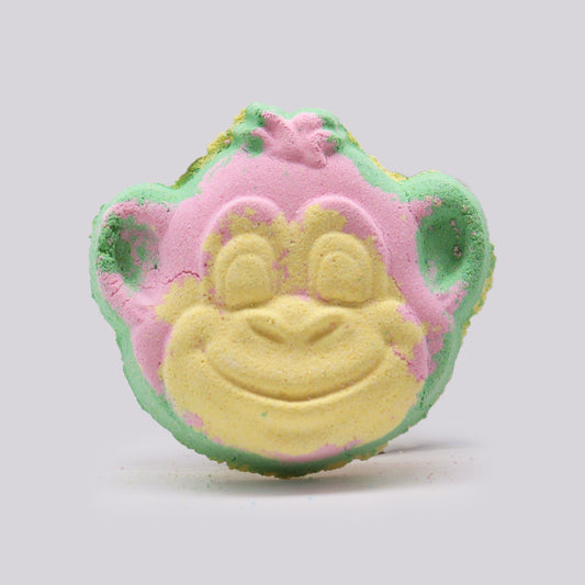 A colorful bath bomb shaped like a monkey face, combining green, pink, and yellow colors, sits against a plain light gray background.