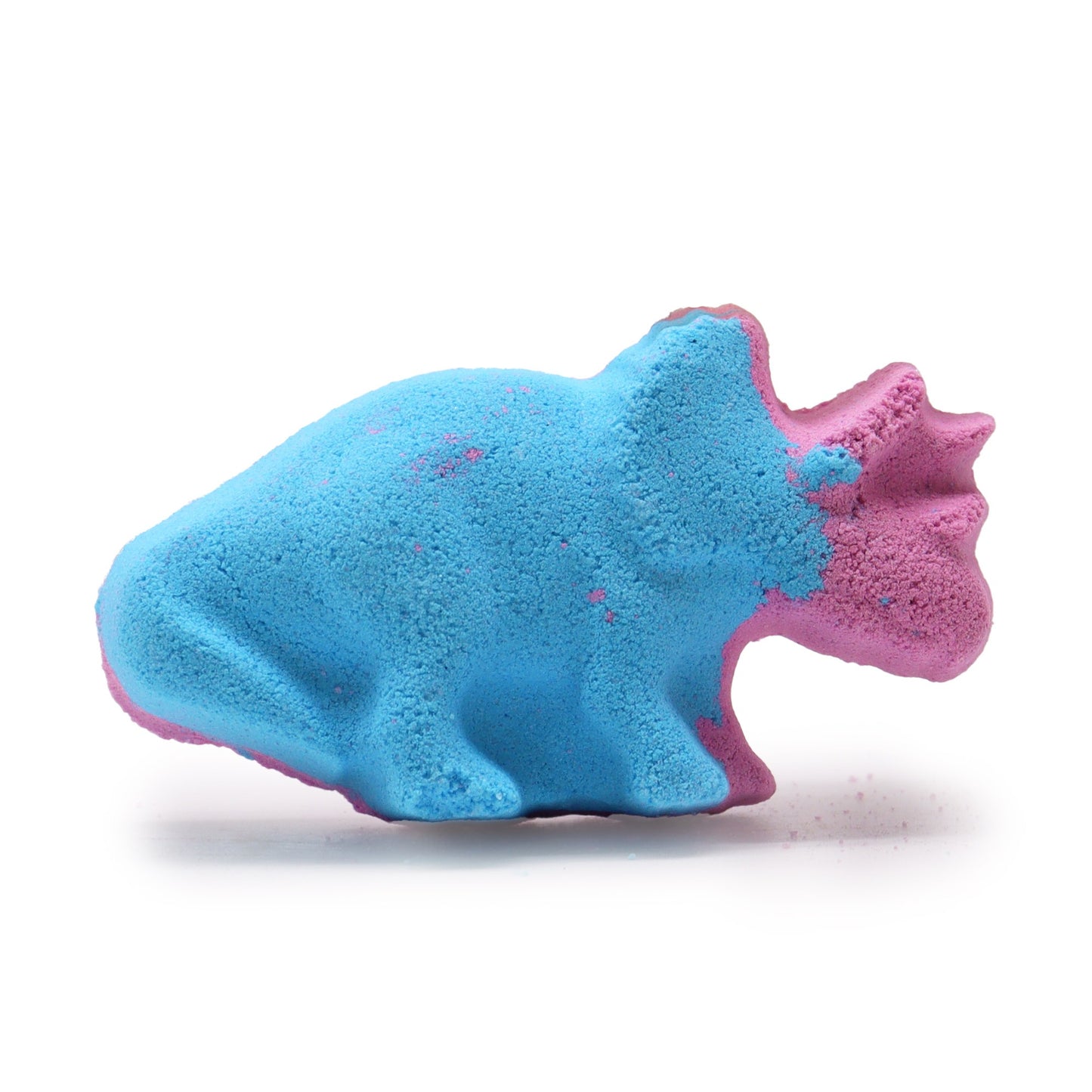 A blue and pink bath bomb shaped like a dinosaur sits on a white surface, appearing textured and slightly powdery in a well-lit environment.