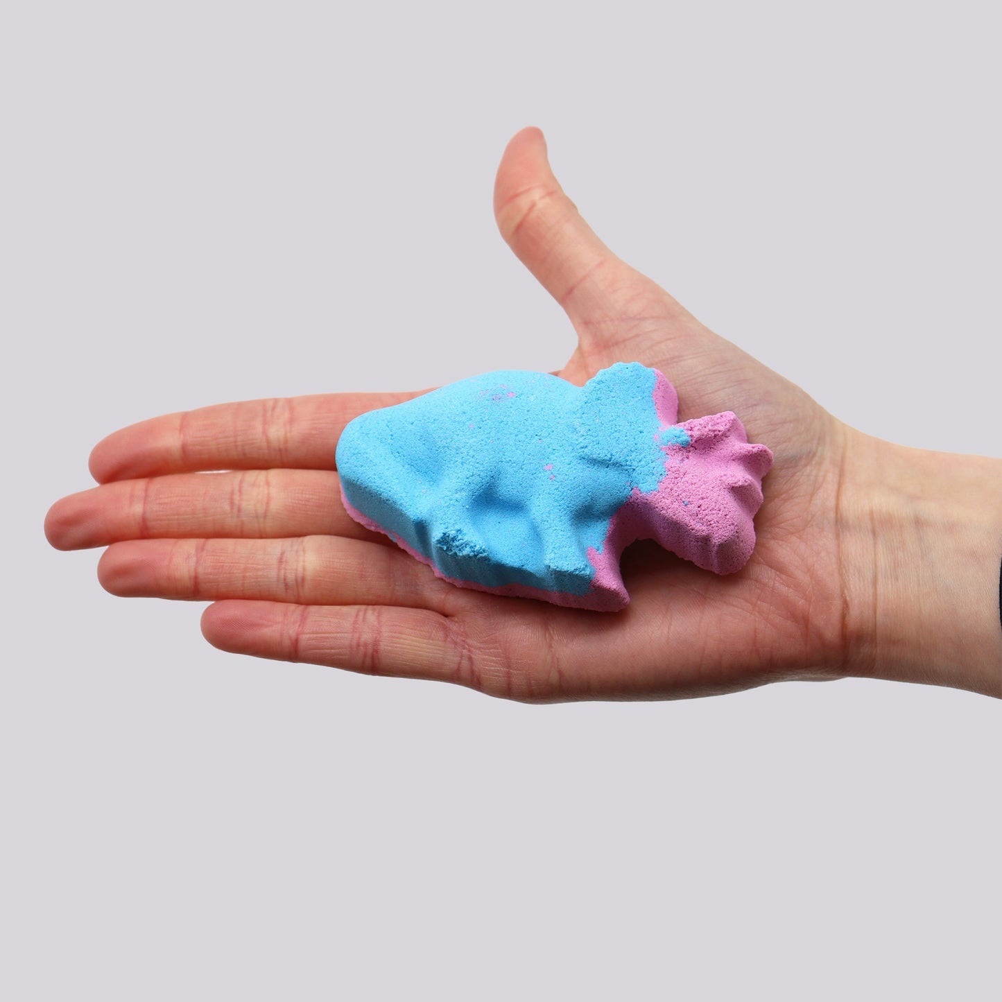 A textured, blue and pink fish-shaped bath bomb rests on an open palm against a plain gray background.