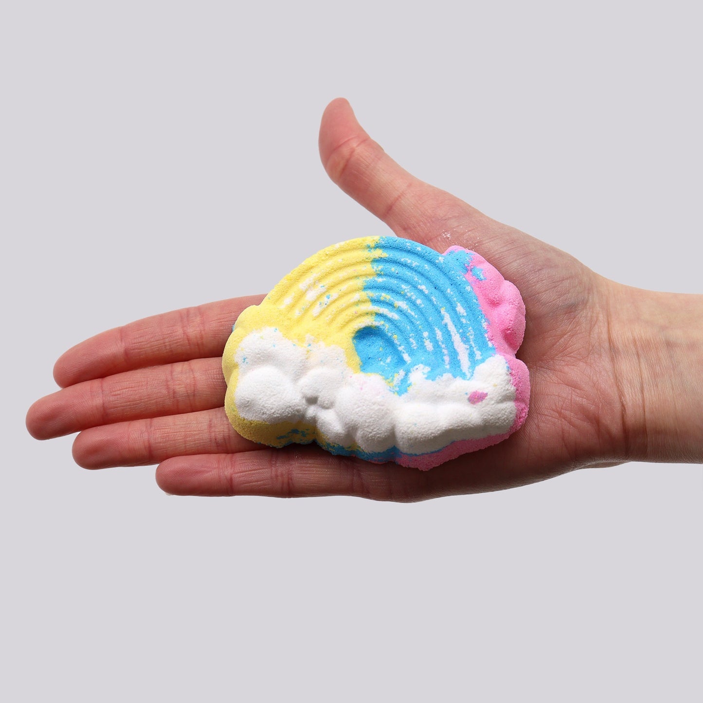 A hand holds a multicolored rainbow-shaped bath bomb with colors of yellow, blue, pink, and white resembling clouds, set against a plain gray background.