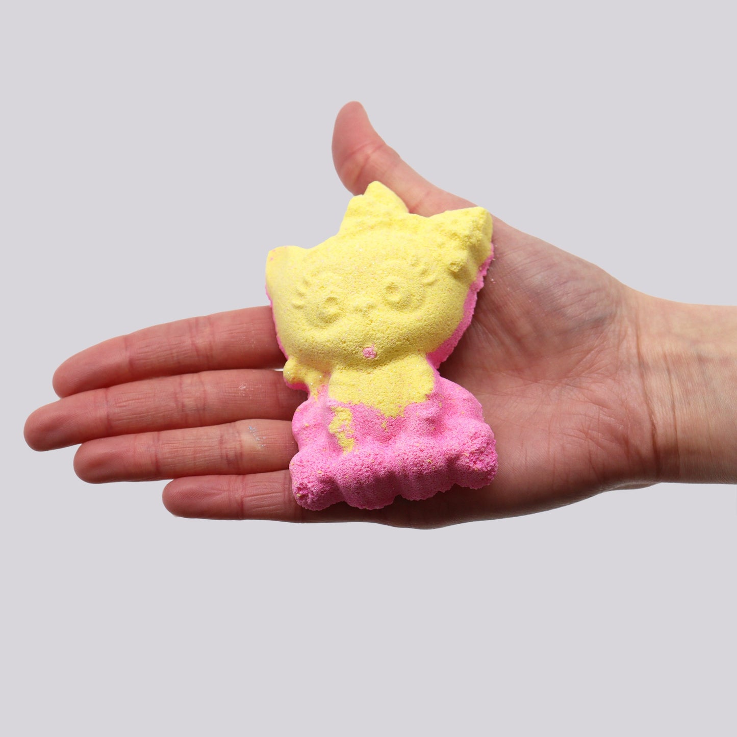 A hand holds a yellow and pink cat-shaped bath bomb, presenting it against a plain, light gray background.
