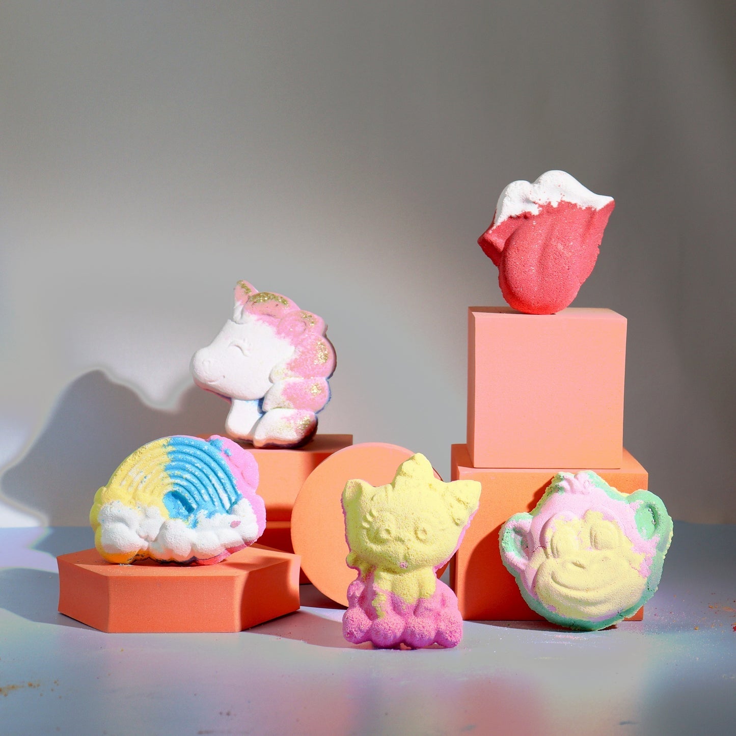 Colorful bath bombs shaped like a unicorn, rainbow, cat, and monkey are displayed on peach-colored geometric platforms in a well-lit setting.