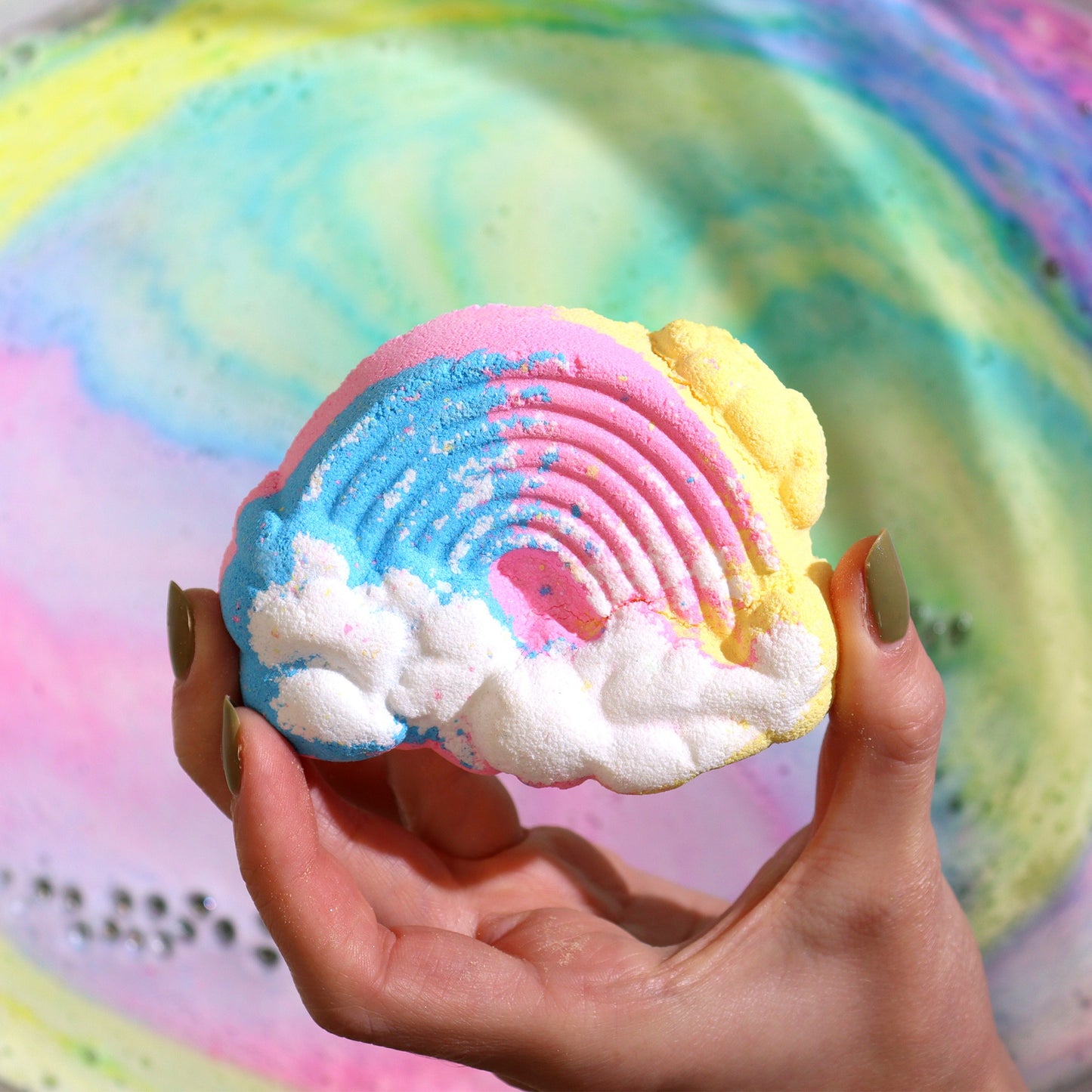 A hand holds a colorful rainbow-shaped bath bomb, featuring pink, blue, and yellow hues with cloud details, above swirling pastel-patterned water.