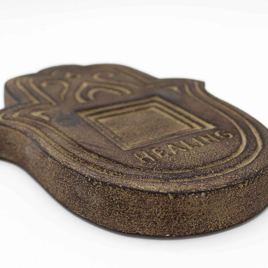 Brown wooden plaque shaped like a hand, featuring intricate engravings and the word "HEALING." It rests on a white surface, suggesting a peaceful, decorative setting.