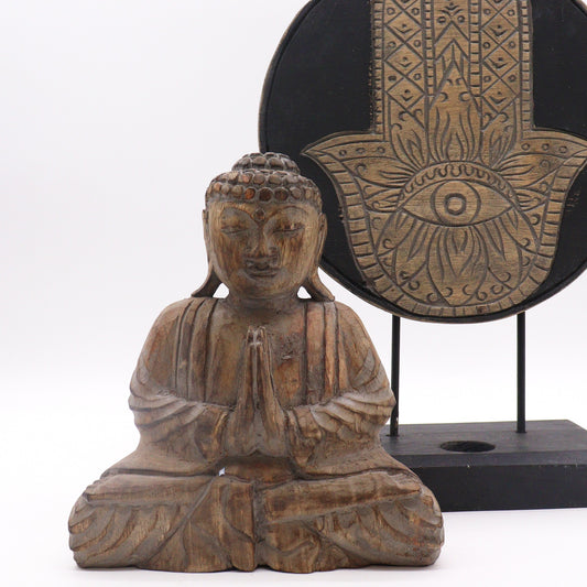 A wooden Buddha statue sits in a meditative pose with hands joined. Behind, a decorative circular plaque features an ornate hamsa design with an eye, displayed on a black stand.