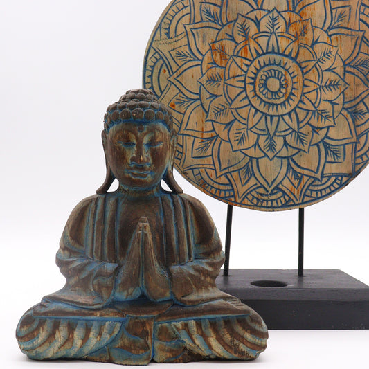A carved Buddha statue sits with hands in prayer, in front of a decorative round mandala design, set on a simple black stand and against a neutral background.