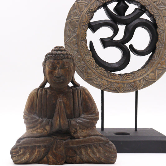 A small wooden Buddha statue sits in a meditative pose with hands in prayer, placed next to a decorative stand featuring a large, carved Om symbol on a black base.