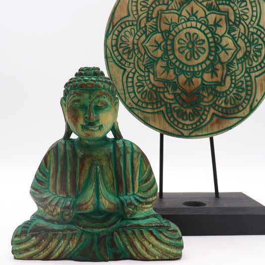 A carved, green Buddha statue sits with palms together in front of a circular wooden mandala on a stand, set against a plain background.