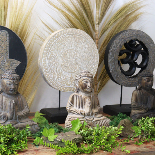 Three Buddha statues, seated with hands in prayer, are placed on a wooden surface surrounded by green foliage. Behind them, circular decorative panels and tall grasses create a serene atmosphere.