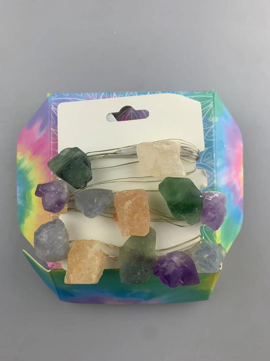 Raw crystals of assorted colors are strung along a thin wire, displayed on a tie-dye patterned cardboard holder against a plain grey background.