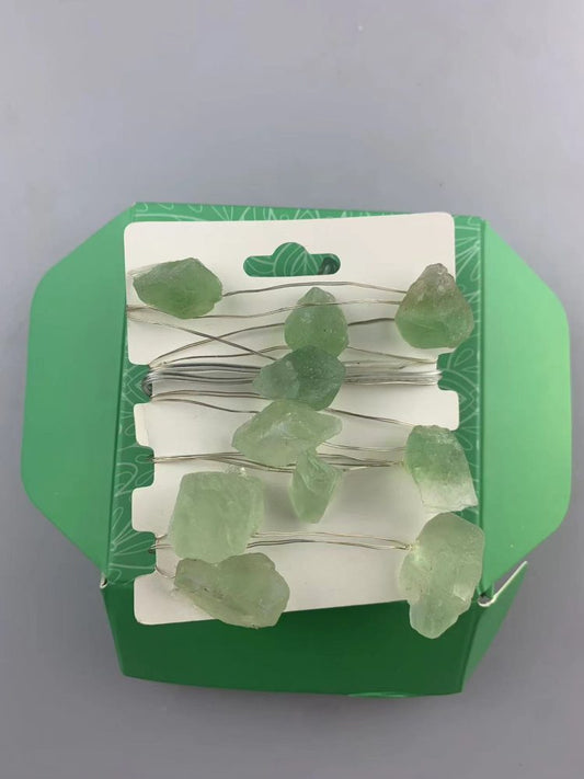 Ten light green rough gems are strung on a silver wire. The wire and gems are attached to a white card that is folded into a green container.