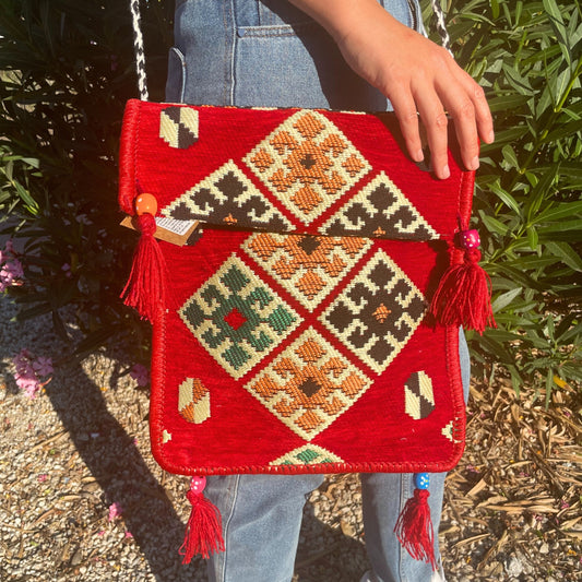A red woven bag with geometric patterns hangs from a shoulder, its flap partially closed by a hand. Tassels adorn the bag. Green foliage is visible in the background.
Text: od Anden, Slovania Co, BL Care Cas, Esparta Comp, 58 1XT LIK, All rights reserved.