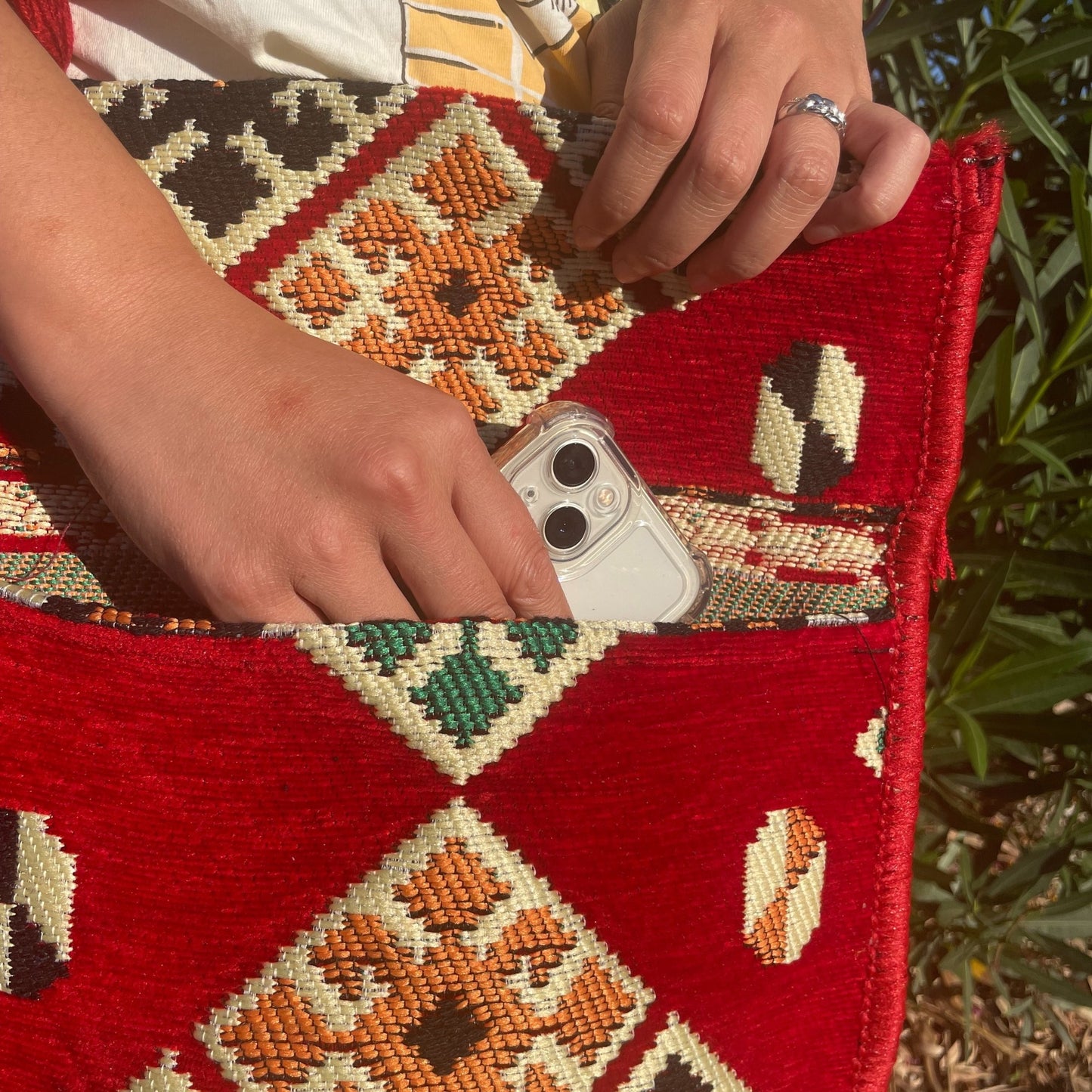 A person inserts a phone into a red woven tote bag with a geometric pattern. This action occurs outdoors, with green foliage visible in the background.