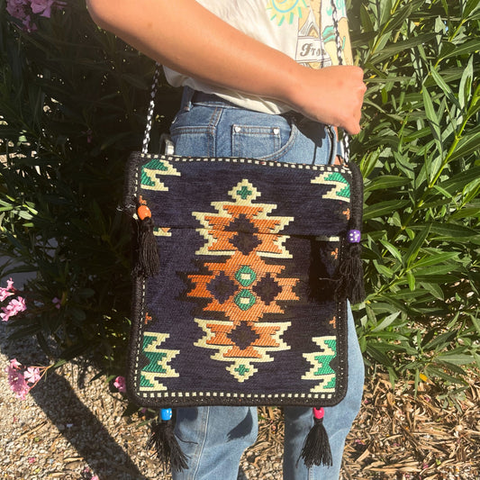 A person wearing jeans has an embroidered crossbody bag hanging from their shoulder. The bag has tassels and geometric patterns in green, orange, and off-white. Pink flowers are in the background.