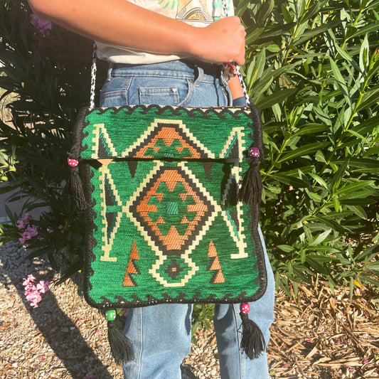 A person holds a green, patterned shoulder bag with tassels. The bag has an orange and black diamond design and a black and white braided strap. The background shows greenery.