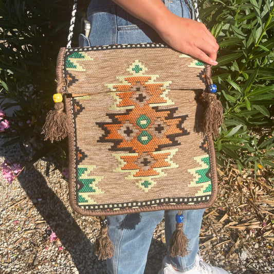 A person holds a large, patterned tapestry bag. The bag features geometric designs in earth tones, with colorful tassels and a braided strap. The person is standing outdoors near foliage.