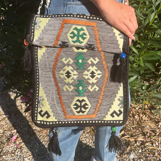 A person holds an ethnic-patterned bag, made of gray fabric, decorated with orange, green, and pale yellow geometric shapes and black tassels, in front of a leafy shrub.