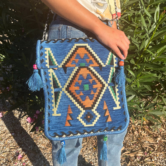 A person holds a blue woven bag with colorful geometric patterns and tassels. The person is wearing jeans and standing outdoors near green foliage and gravel ground.