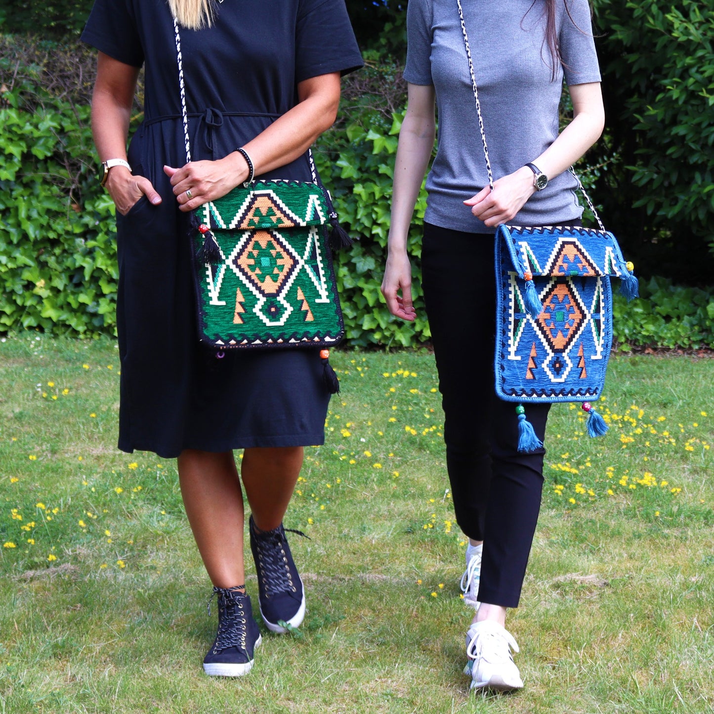 Two women walk side-by-side, each wearing a colorful, patterned, tasseled cross-body bag. They are on a green lawn sprinkled with small yellow flowers, with a green bush behind them.