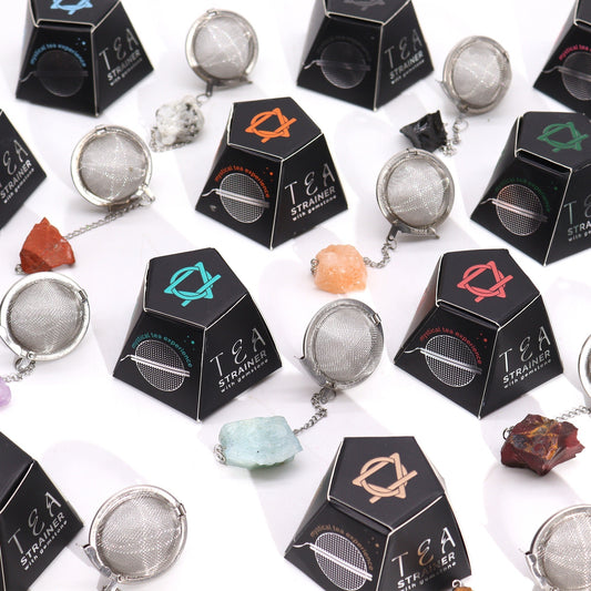 Round metal tea strainers are attached to various crystals, each packaged in black boxes labeled "TEA STRAINER, mystical tea experience, with gemstones," arranged on a white surface.