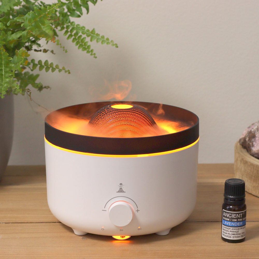 Aroma diffuser emits vapor from a central dome, atop a white base with a dial. Nearby is a bottle of "ANCIENT WISDOM LAVENDER" oil. All is set on a wooden surface, near a plant.