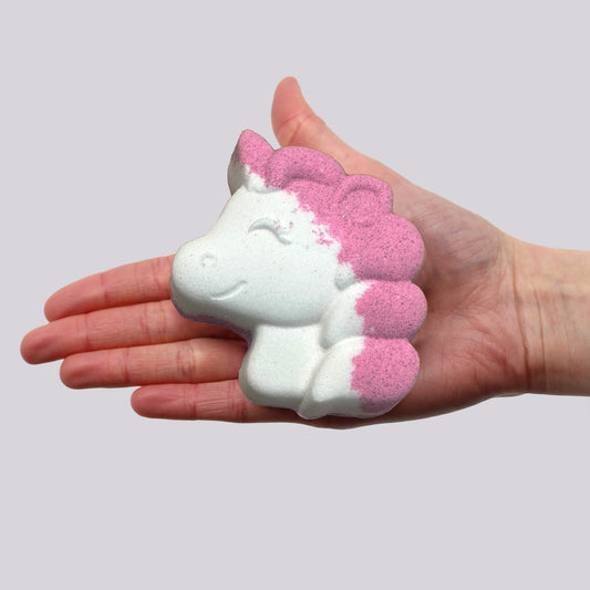 A hand holds a unicorn-shaped bath bomb featuring pink and white colors, depicted against a plain, grey background.