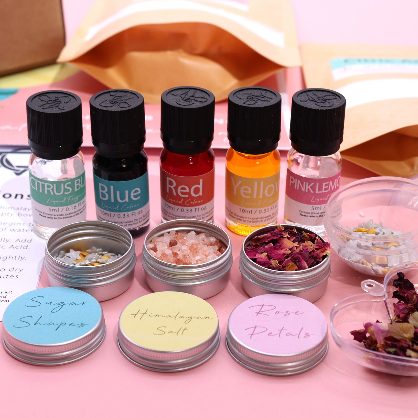 Bottles of liquid colors labeled Citrus Blue, Blue, Red, Yellow, Pink Lemon. Metal tins holding sugar shapes, Himalayan salt, rose petals. Background includes open envelopes on a pink surface.