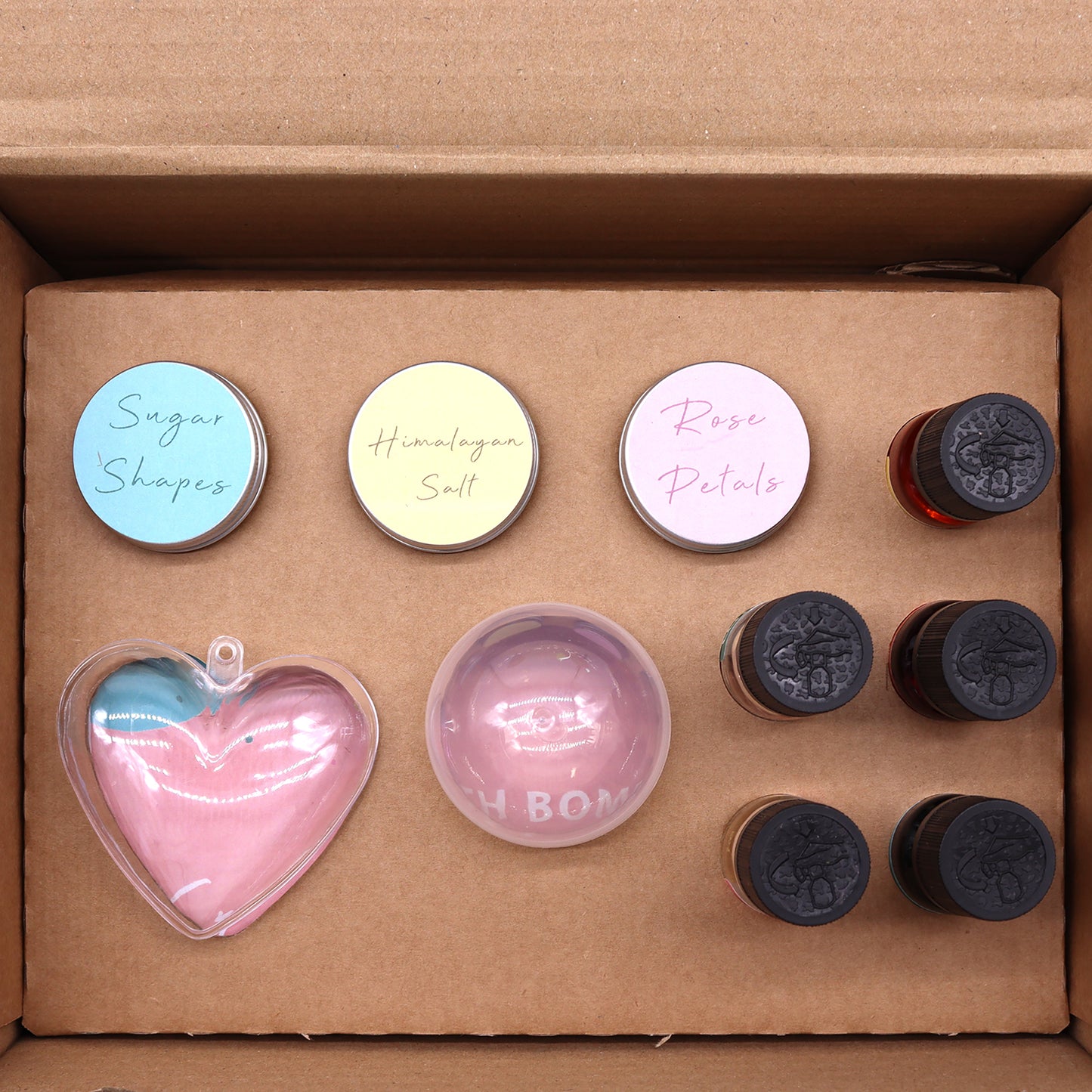 Box containing three small tins labeled "Sugar Shapes," "Himalayan Salt," "Rose Petals," a heart-shaped item, a round item marked "H.BOM," and six small bottles, arranged neatly inside.