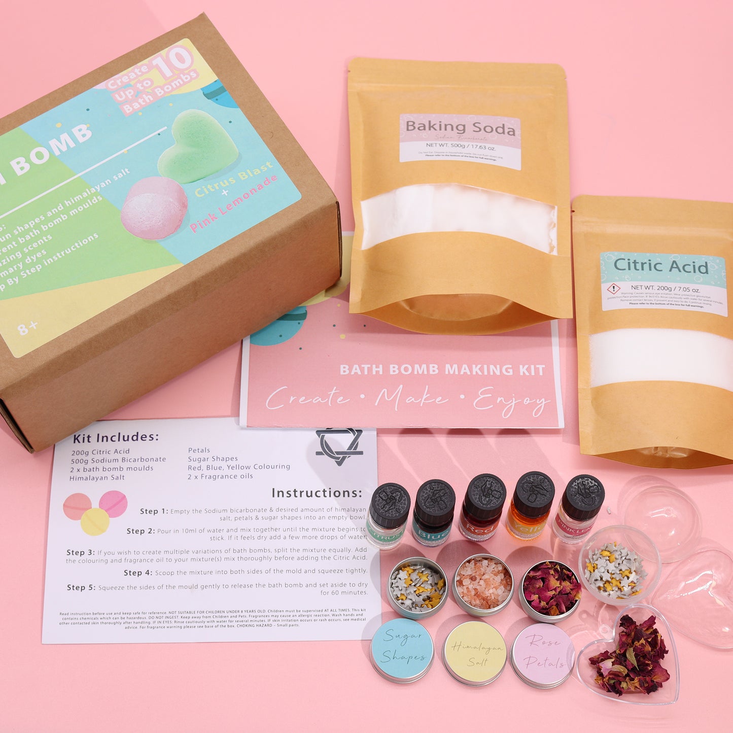 A bath bomb making kit displayed on a pink surface includes baking soda, citric acid, fragrances, molds, petals, and colored powders. Text on packaging: "BATH BOMB MAKING KIT - Create, Make, Enjoy." Instructions detail mixing steps.