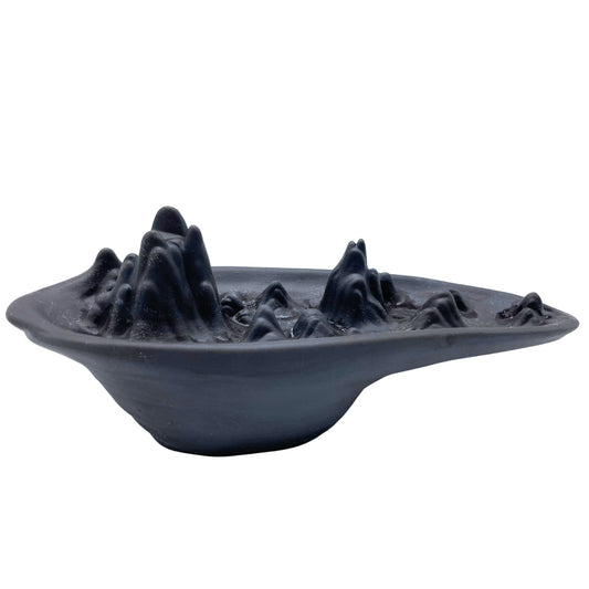 A dark, ceramic bowl features a sculpted mountain range inside, creating a miniature, artistic landscape. The mountains rise sharply from the smooth surface of the bowl.