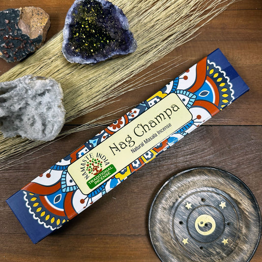 A colorful box labeled "Nag Champa Natural Masala Incense" rests on a wooden surface, accompanied by geode stones, dry grass, and a round carved incense holder featuring a yin-yang design.