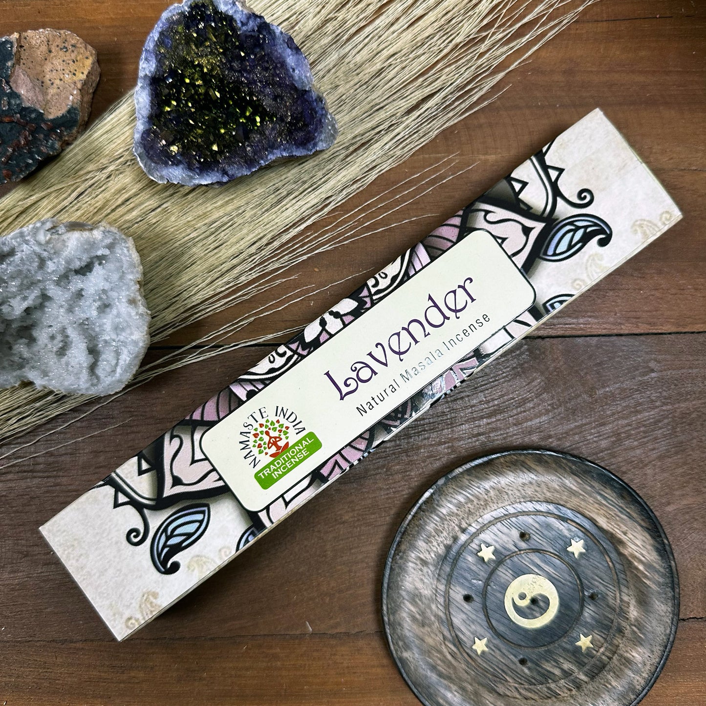 Incense package labeled "Lavender Natural Masala Incense" with Namaste Indian logo, rests on wood beside amethyst and other crystals, dried grass, and a round incense holder featuring a yin-yang symbol and stars.