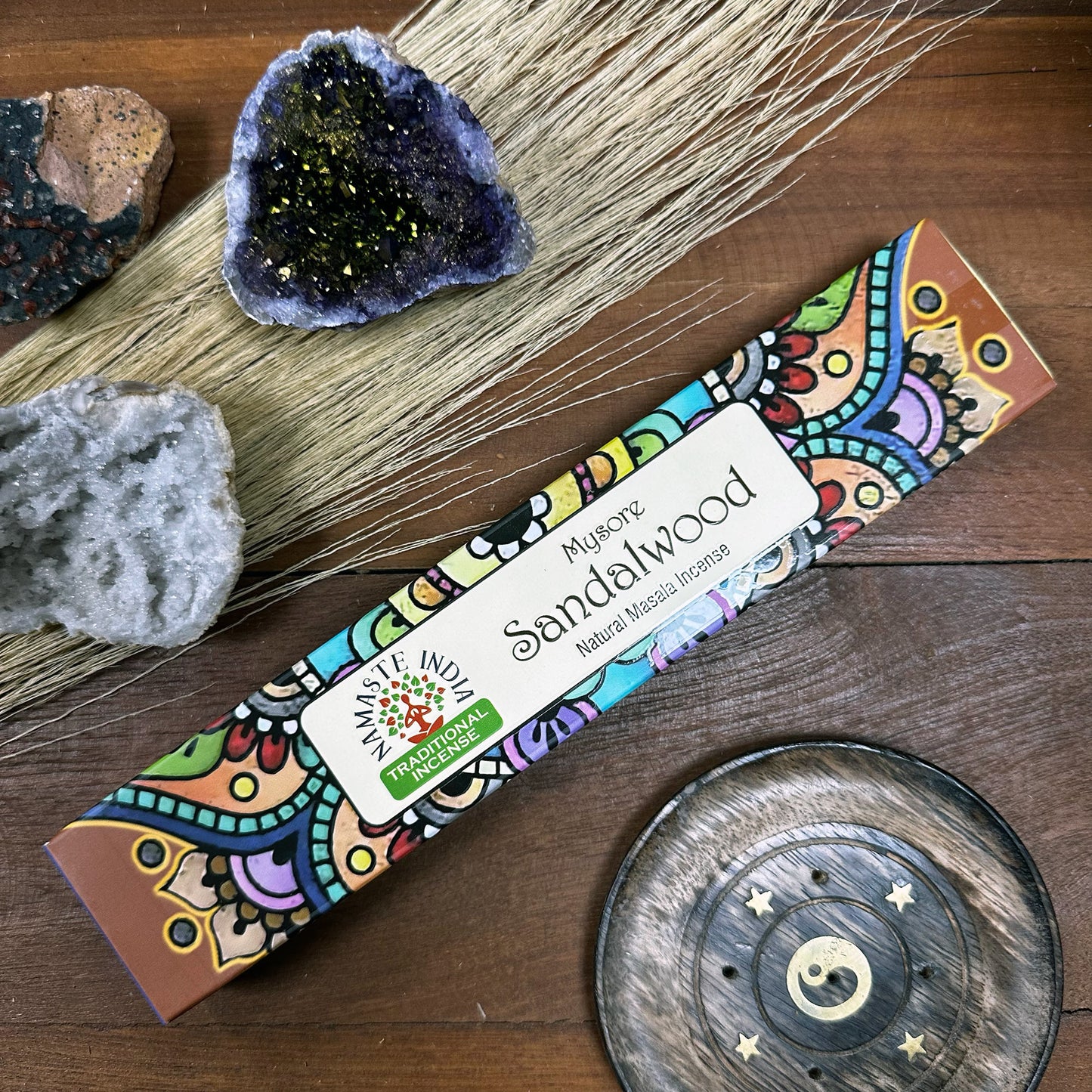 Incense pack labeled "Mysore Sandalwood, Natural Masala Incense," rests on a wooden surface with colorful designs. Nearby are crystals, wheat stalks, and a decorative wooden dish featuring a yin-yang symbol.