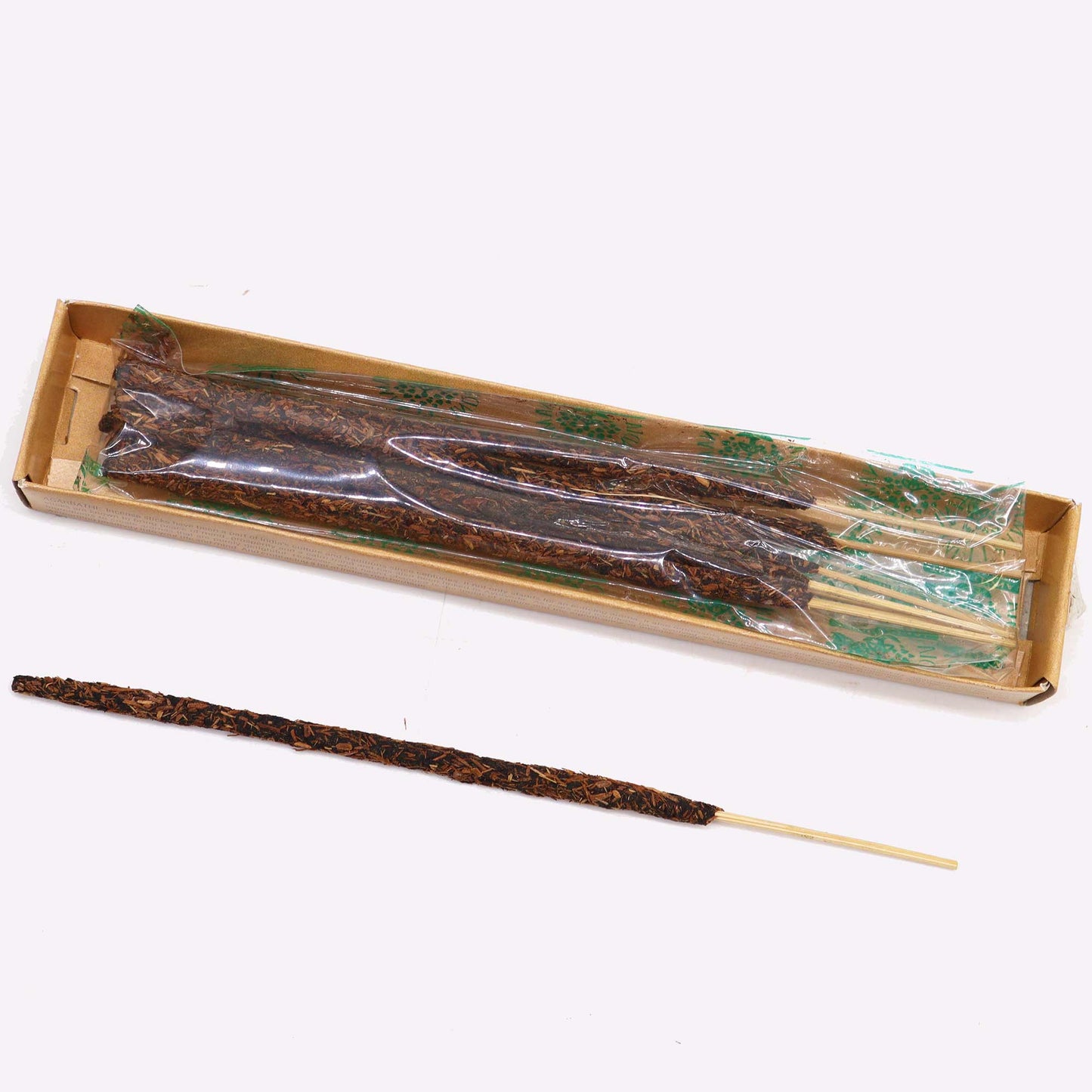 A bundle of long, brown, twig-like skewers wrapped in plastic sits in a cardboard box. One skewer lies outside the box on a light surface.