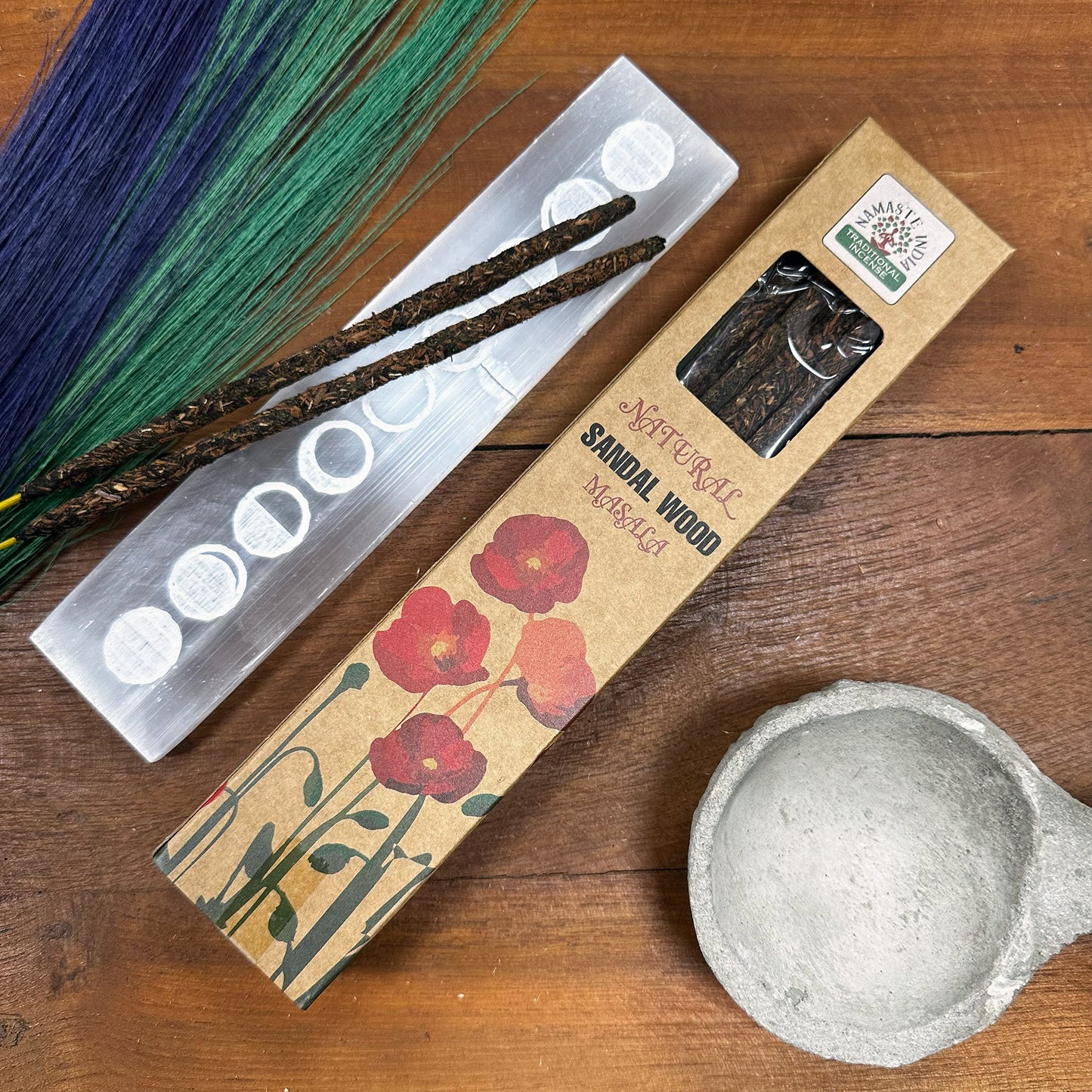 Incense sticks rest on a decorative holder beside a box labeled "Natural Sandalwood Masala" adorned with red poppies. A round, gray stone holder sits nearby on a wooden surface.