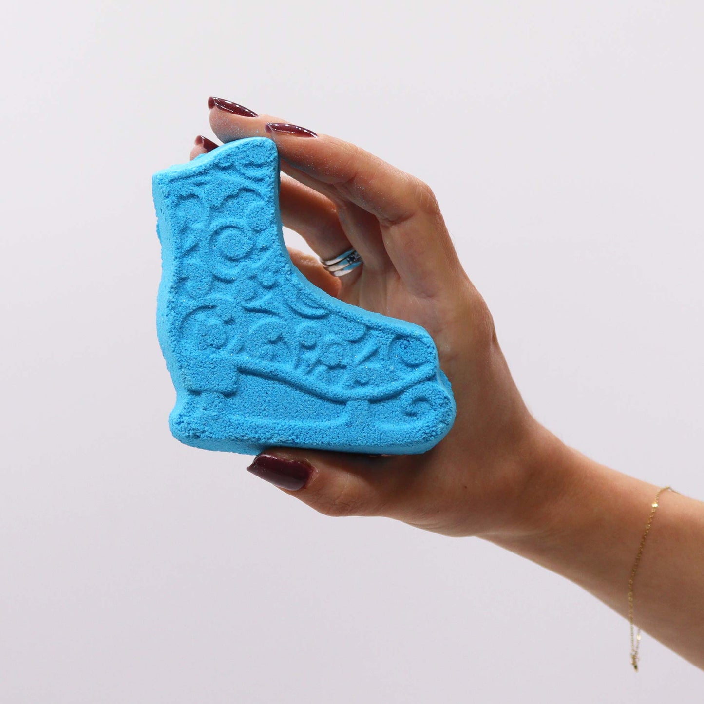 A hand with maroon nails holds a blue, roller skate-shaped bath bomb, featuring intricate swirl designs, against a plain white background.
