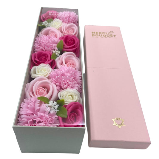 A long, rectangular box containing an arrangement of pink and white soap roses and carnations sits next to its matching lid. MERCIE BOUQUET Floral Celebrations is printed on the lid.