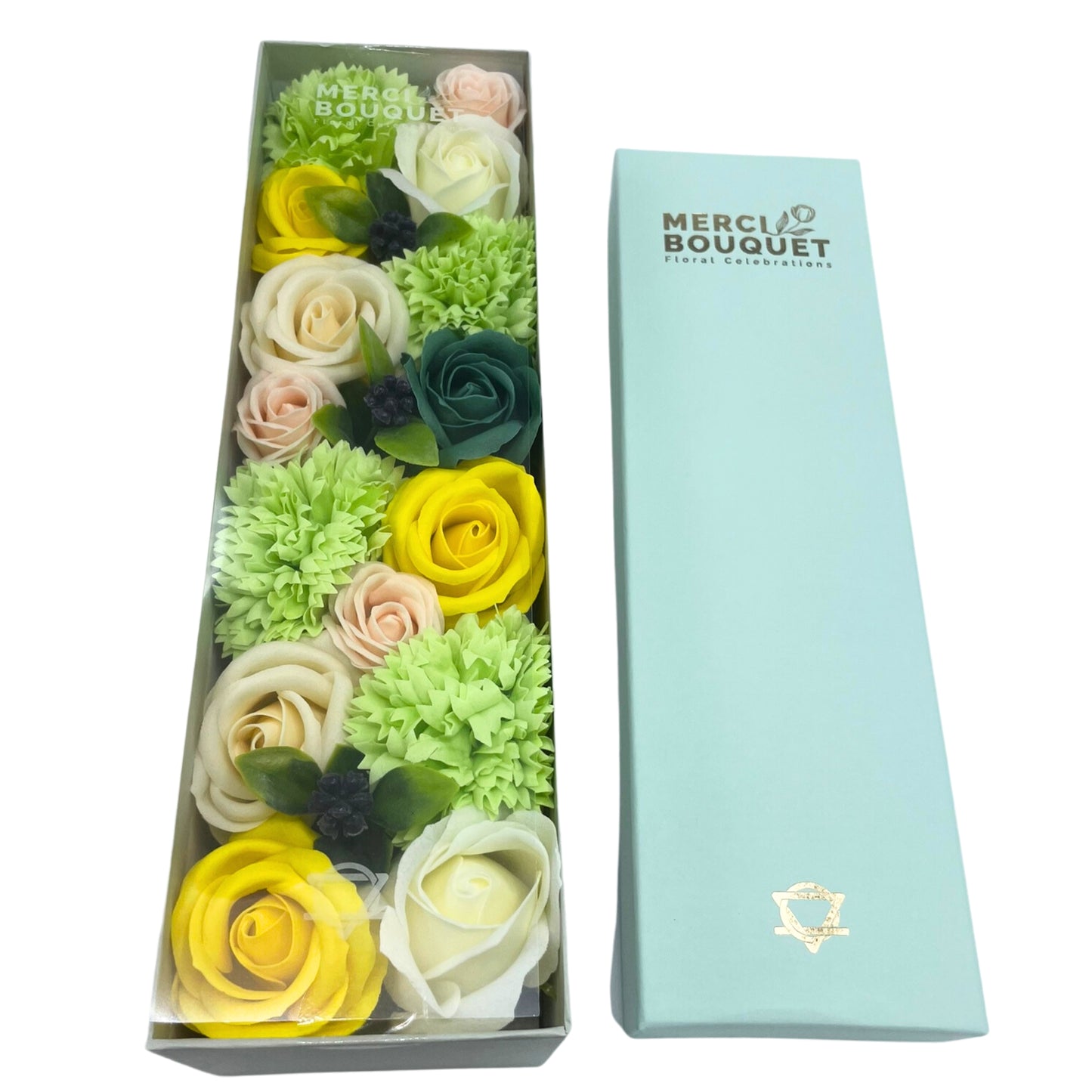 A soap flower bouquet, arranged in a clear box, sits beside its light-blue box. The box displays "MERCI BOUQUET Floral Celebrations".