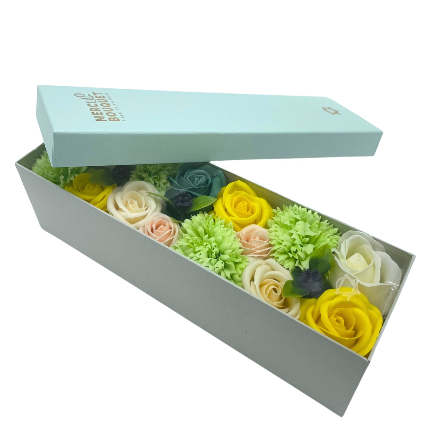 A light-grey box containing a bouquet of pastel and yellow soap roses and carnations rests on a white background. The lid, a light-blue color, is ajar. The box is labeled “MERCIELO BOUQUET Floral Candles”.
