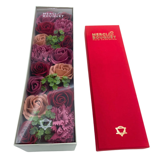 A box of soap roses and carnations in varying shades of red and pink sits beside a red box. Both boxes display the logo "MERCI BOUQUET Floral Celebrations".